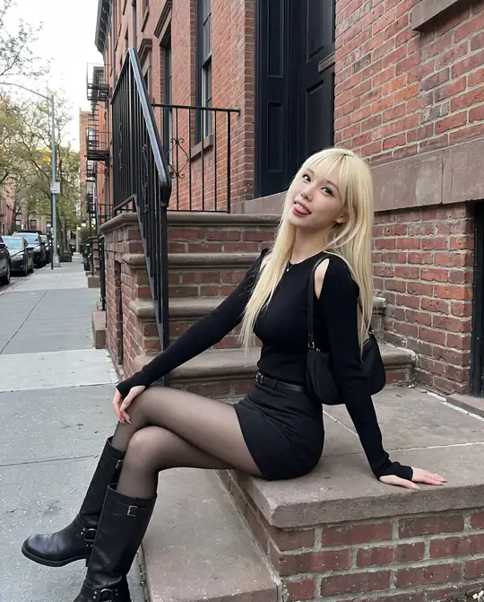 A blonde person in a black shoulder-cutout top, black miniskirt, sheer tights, and buckled boots sits on brick steps.