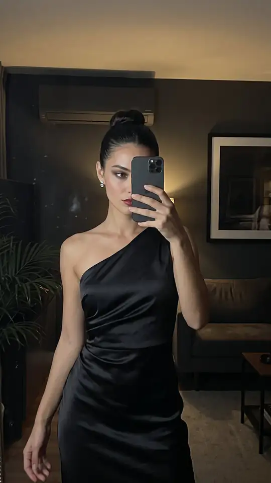 A woman in a black one-shoulder satin dress takes a mirror selfie in a dim room.