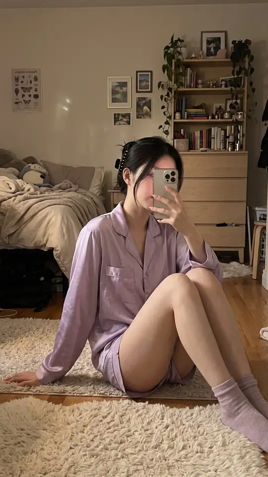 A woman in lavender pajamas takes a mirror selfie while sitting on a bedroom rug.