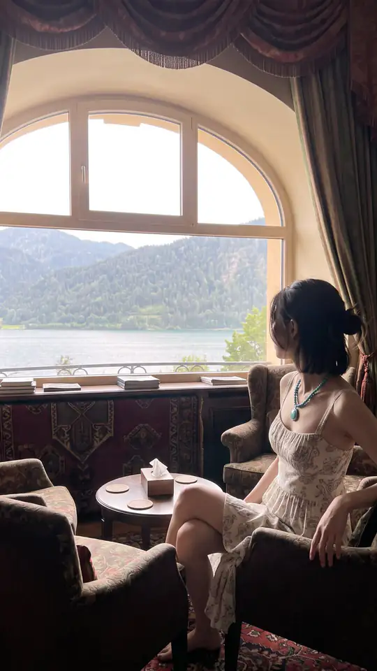 A woman in a patterned dress sits in an armchair beside a large window overlooking a lake.