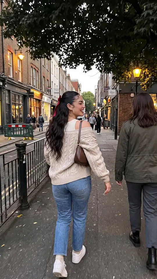 A woman in a cream cable-knit cardigan and jeans looks back while walking along a shop-lined street.