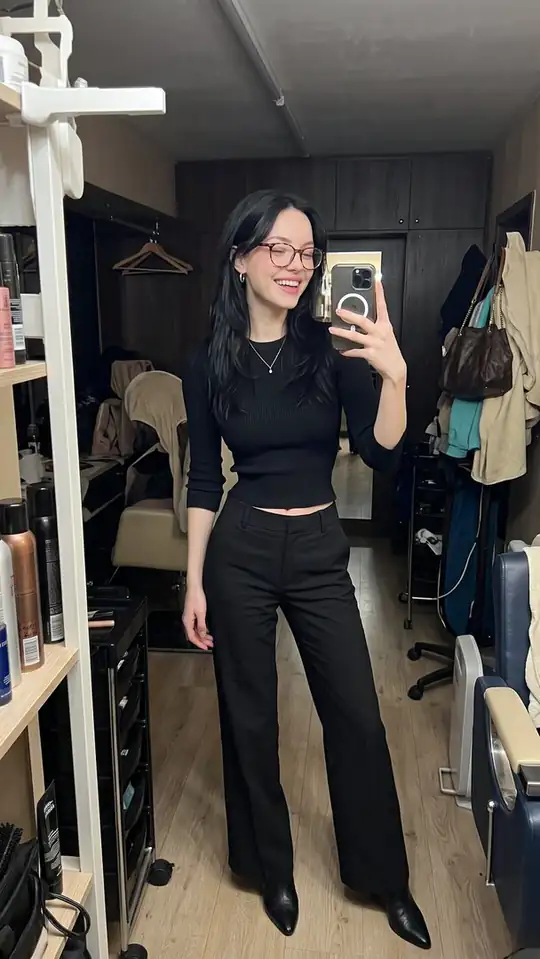 A woman wearing glasses, a black cropped top, black trousers and boots takes a salon mirror selfie.